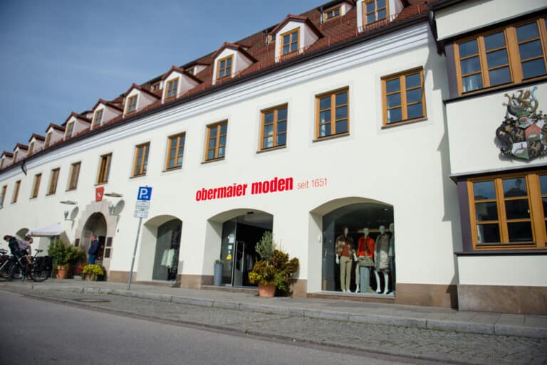 weiße mehrstöckige Fassade mit roten Dachgauben, gelb gerahmte Fenster, roter Schriftzug obermaier moden seit 1651, Auslage mit Modefiguren, Straßenszene mit Bäumen und Fahrrädern