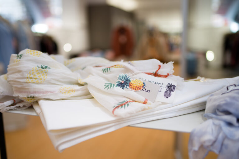 Weiße Tücher mit gelbem Ananas-Motiv liegen auf einem Tisch in einem Modegeschäft von Obermaier Moden; unscharfer Hintergrund mit Kleidung und Regalen.