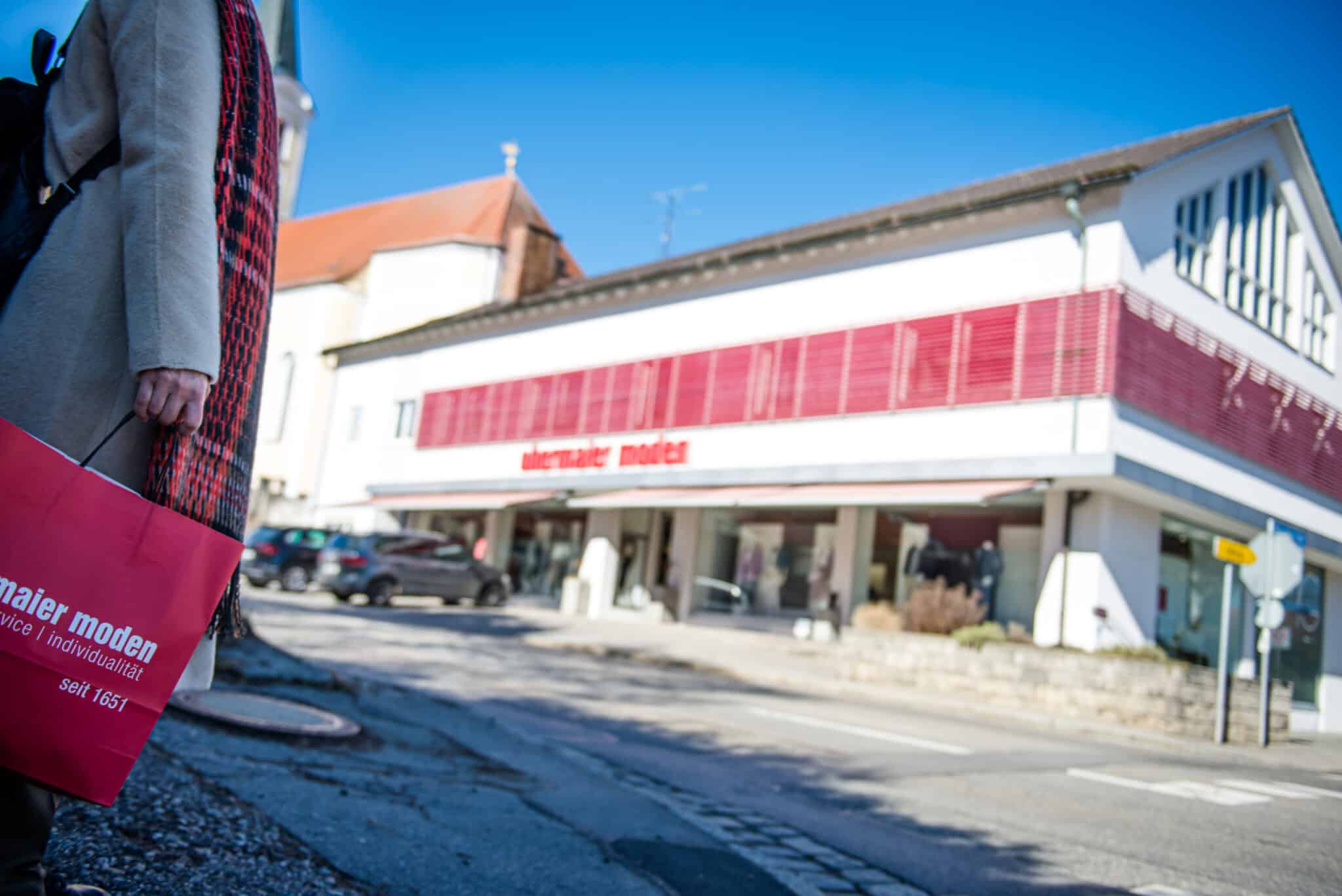 Person mit pinker Einkaufstasche von Obermaier Moden vor dem Stammhaus in Glonn, blauer Himmel, modernes Geschäft mit rotem Fensterladen