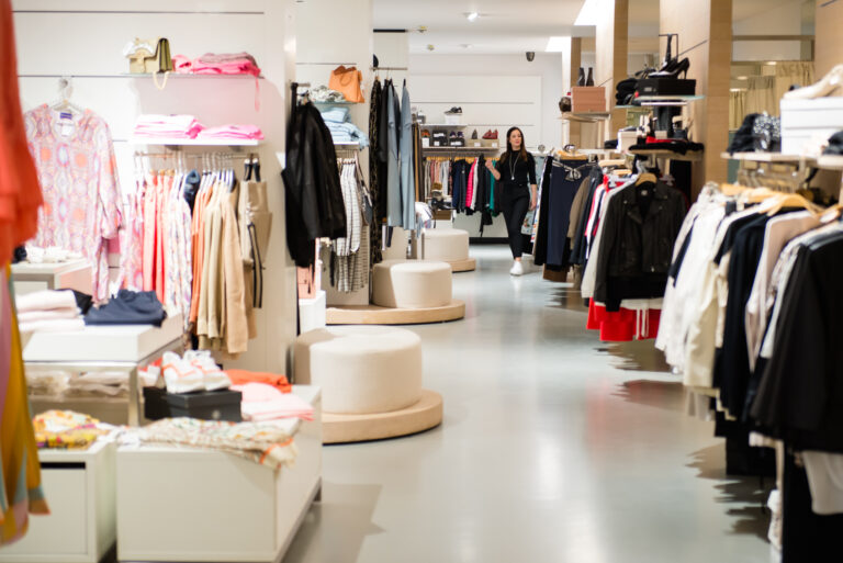Heller Ladenbereich von Obermaier Moden mit Kleiderstangen, farbiger Kleidung und bequemen Sitzgelegenheiten; im Hintergrund eine Person in schwarzer Kleidung bewegt sich durch den Store, Marken Drykorn und Yaya sichtbar.
