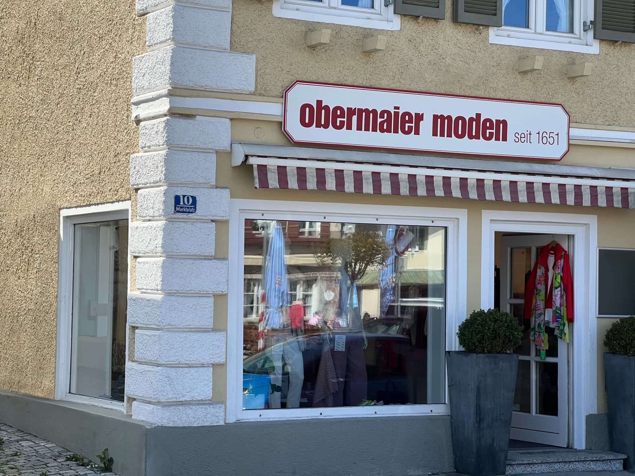Außenansicht eines Modegeschäfts Obermaier Moden mit weißer Fassade und roter Schrift 'obermaier moden seit 1651', großes Schaufenster, rote und weiße Markise, Pflanzkübel neben der Eingangstüre