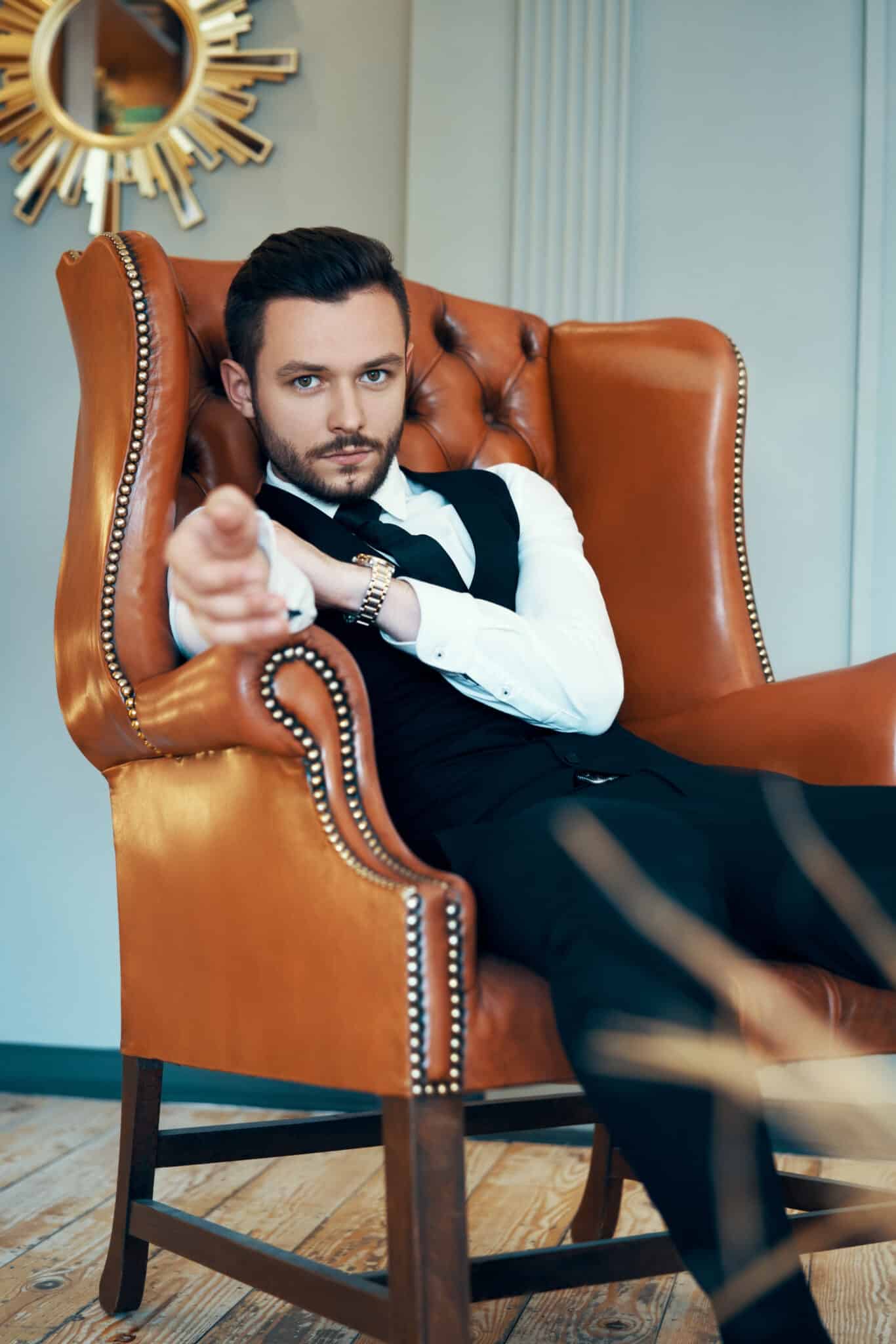 M&auml;nnliche Figur in dunklem Anzug lehnt sich entspannt in einem braunen Leder-Armchair vor modernem Interieur; Marken wie Drykorn, Windsor und Luis Trenker sind Teil des Kontextes der Herrenmode.