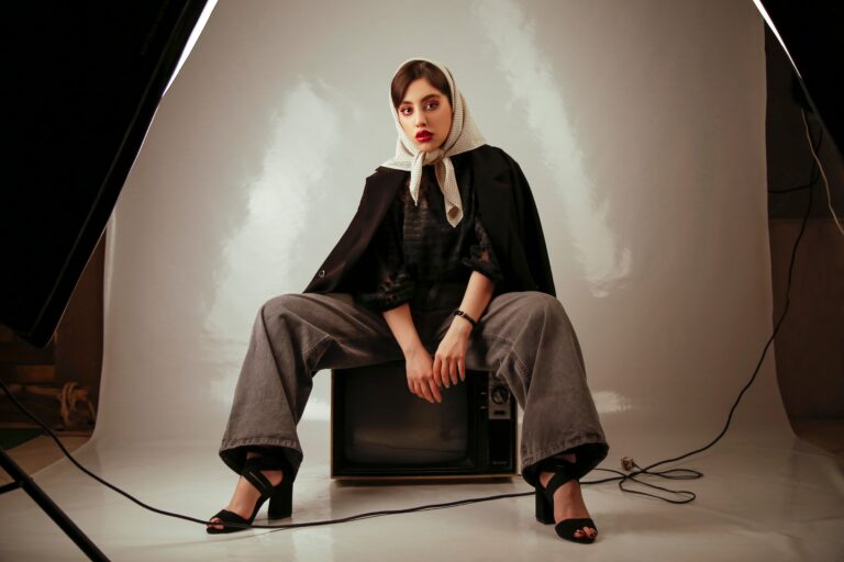 Mamad0vich-Damenlook: Modell sitzt in Studio auf einem alten Fernseher, schwarzer Cape, graue Jeans, beigefarbener Tupfen-Schal um den Kopf; künstlerische Modeaufnahme.