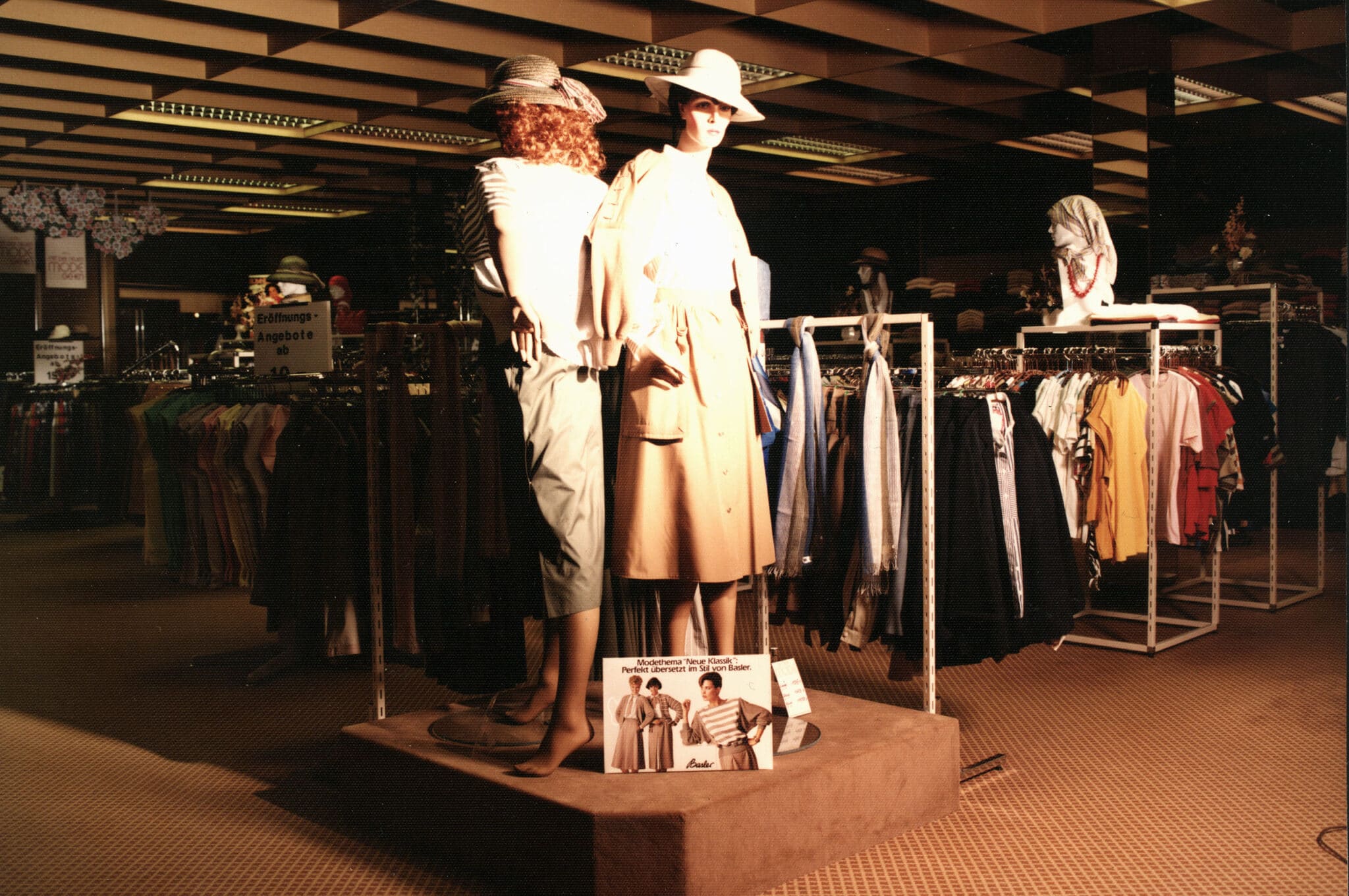 Vintage Modenabteilung in Glonn: zwei auf einem Podest stehende mannequinen tragen Hüte und elegante Kleidung; Kleiderstangen, Warenträger und retro Beleuchtung prägen das Kaufhaus-Setting.